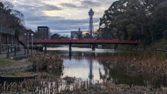 Image de la tour Tsutenkaku se reflétant dans l'eau, avec les bâtiments environnants et un ciel clair, créant une scène urbaine emblématique d'Osaka